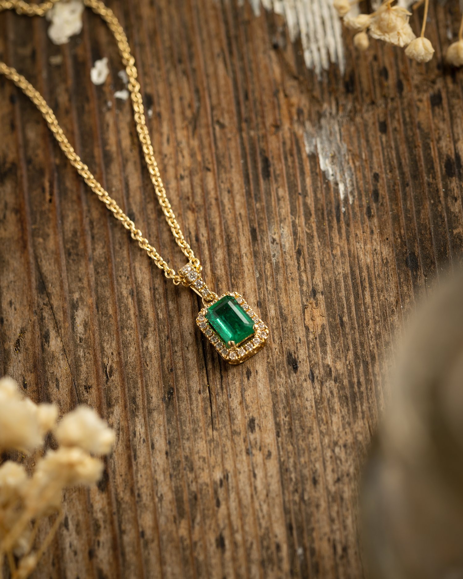 Octagonal Cut Emerald Pendant set within a diamond halo in 18ct yellow gold. Photographed on a wooden background with accents of flowers