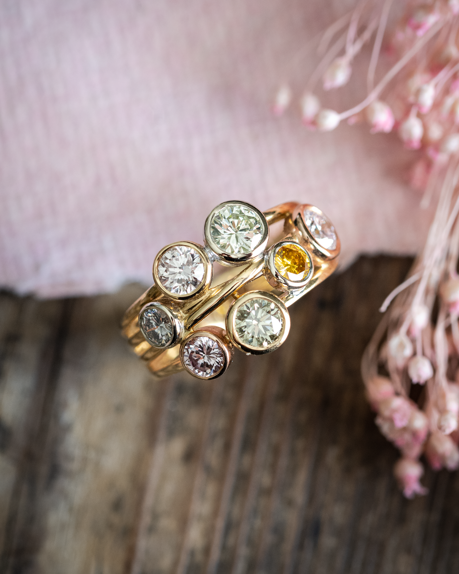 A multi colour sapphire boodles style ring complete with pink, green, white, yellow sapphires photographed on a wooden background with pink paper and pink flowers
