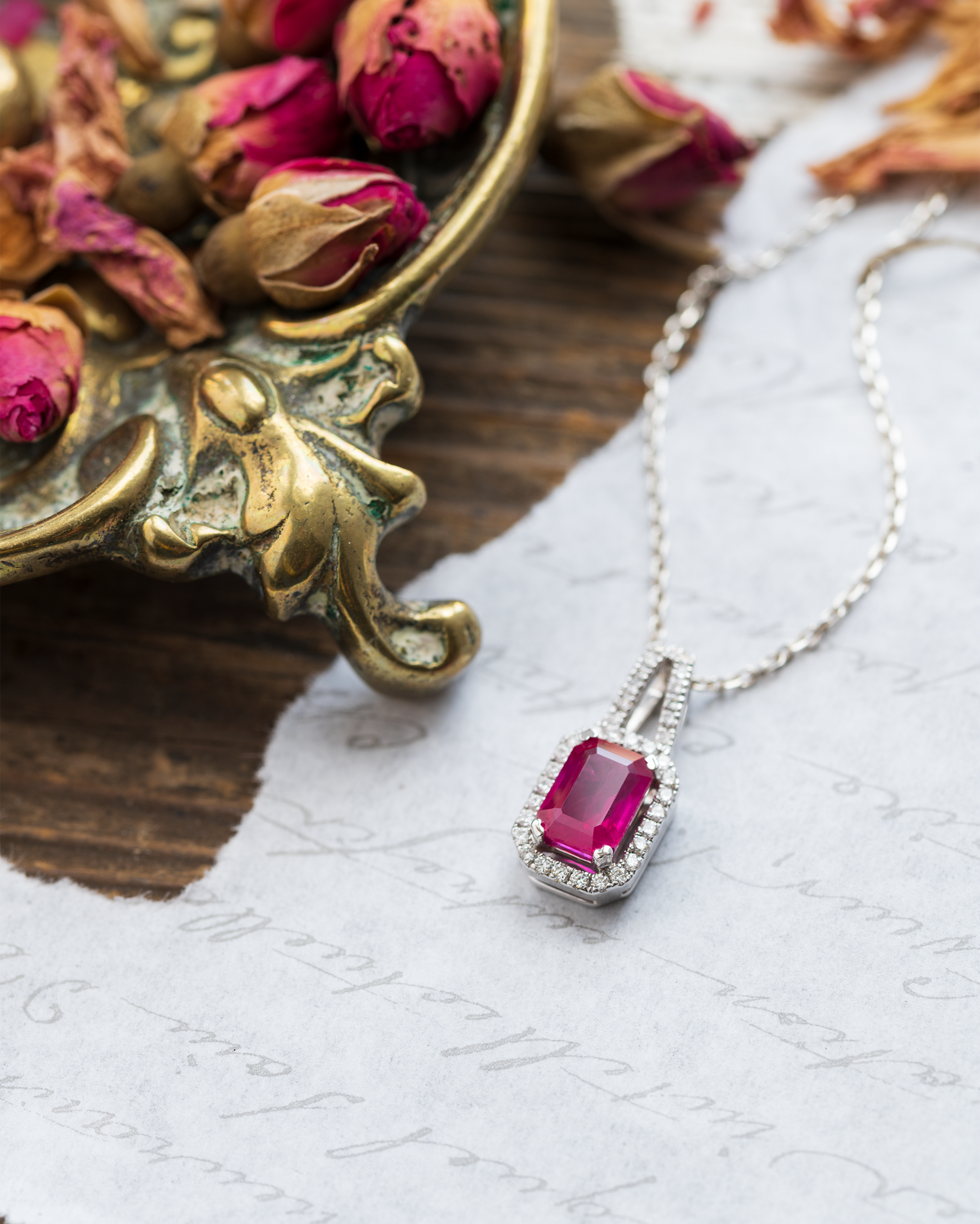 A Ruby and Diamond Pendant photographed on some vintage paper and accompanied with small dried red rose flower heads