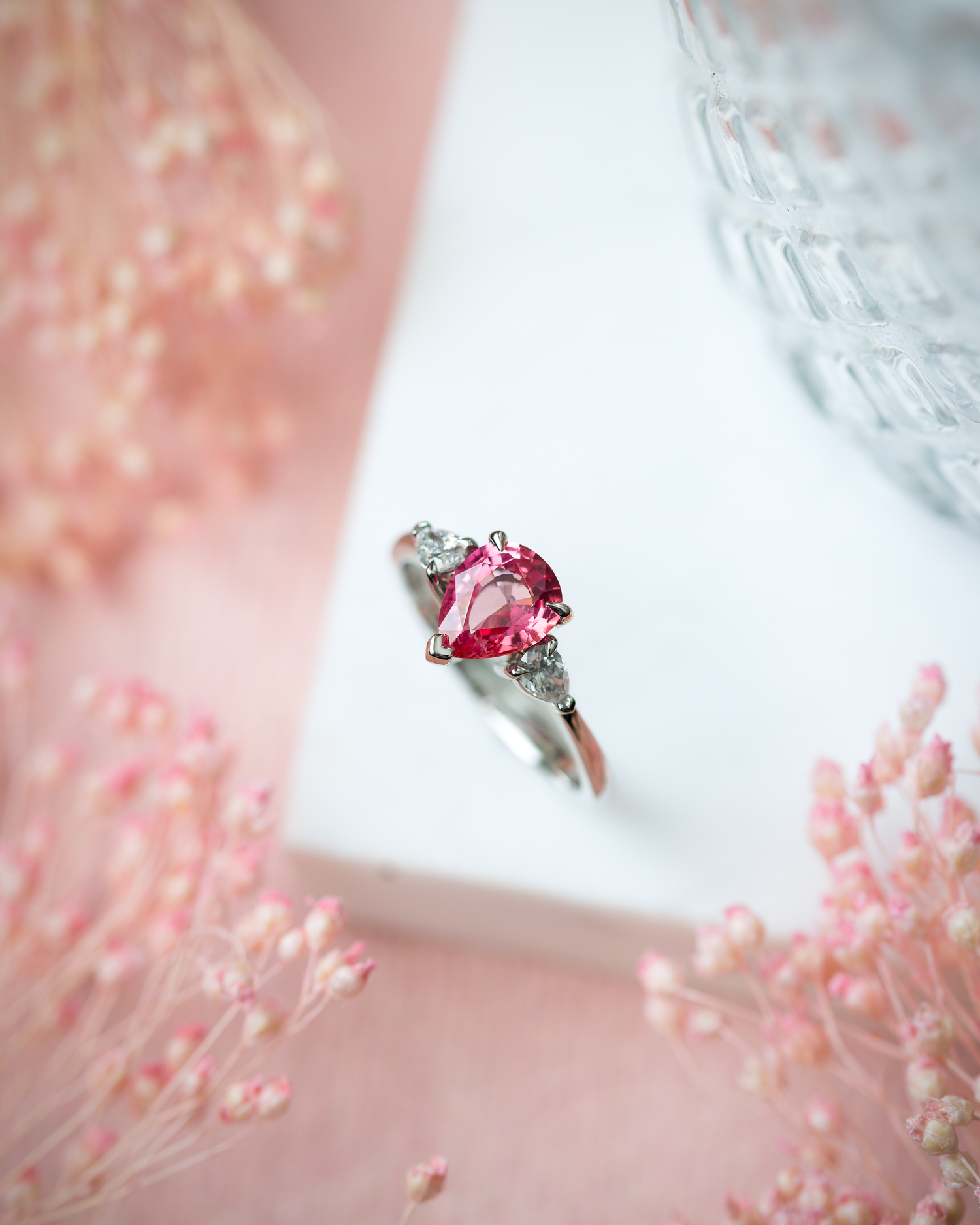 Pear Shape Pink Sapphire Solitaire with pear shaped shoulders, photographed on a marble top with pink accents and pink flowers