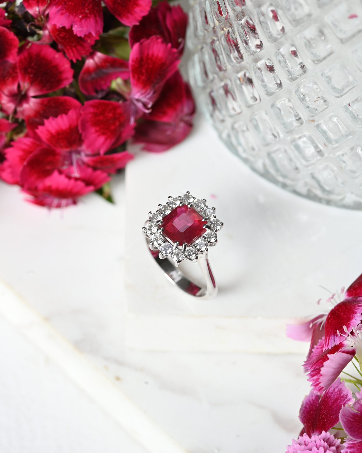 Ruby Solitaire with a Diamond halo, photographed on a white background with pink flowers and a textured vase