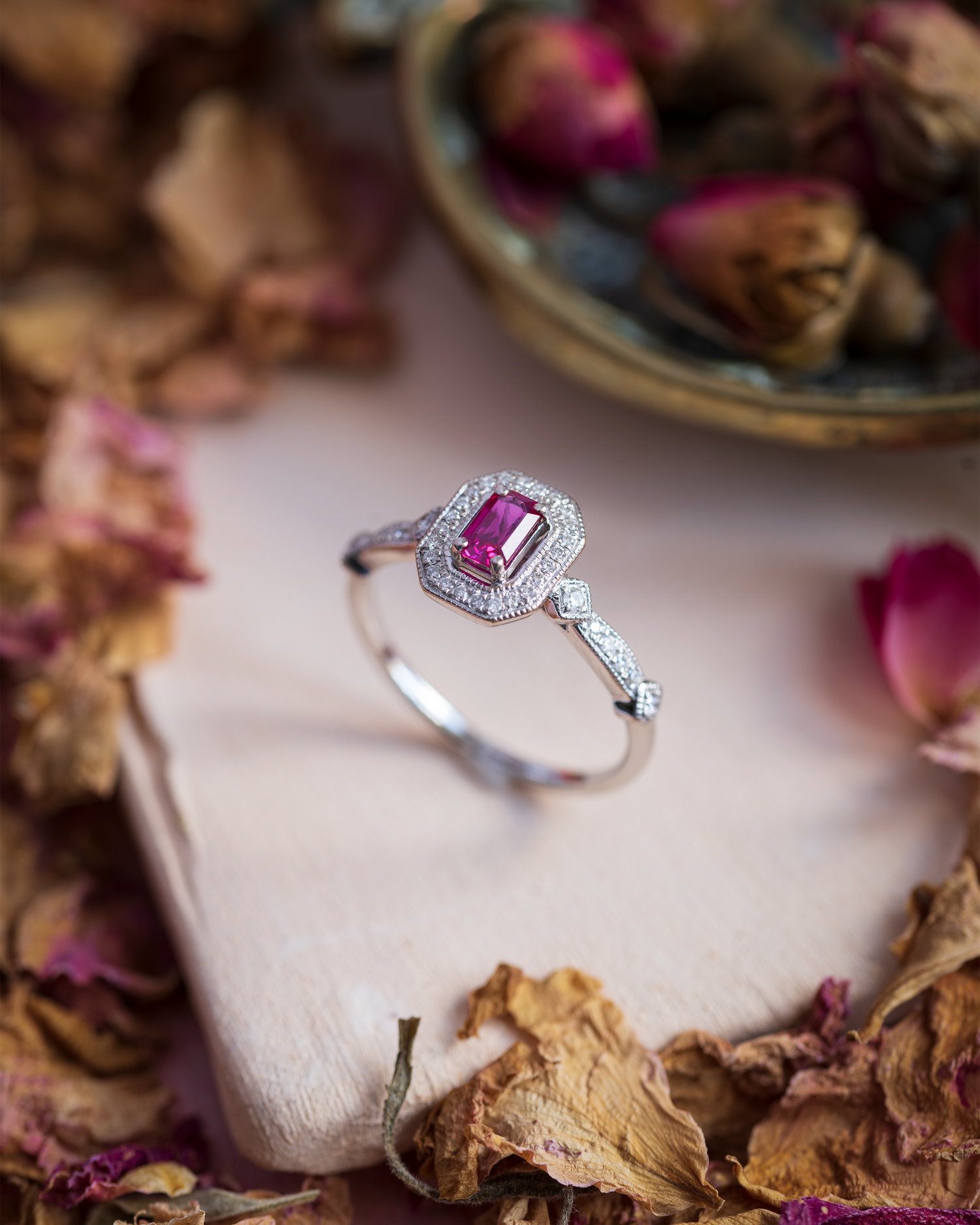 a octagonal cut ruby set within a diamond halo complete with diamond set shoulders, photographed on a hand made tile with dried petals
