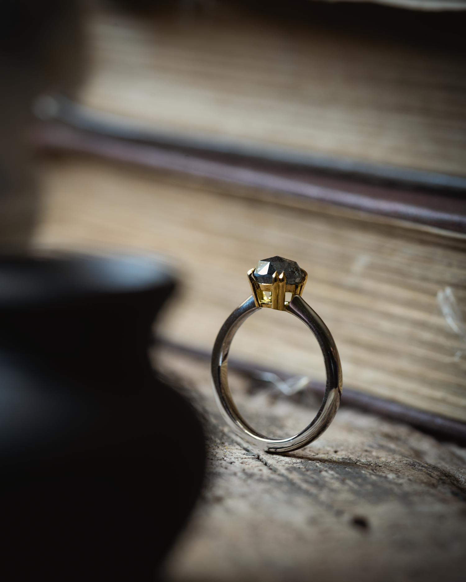 A salt and pepper diamond ring from the side, set in platinum and 18ct with double talon claw setting, photographed on a wooden board with a stack of books behind the ring and a black clay vase in the forefront