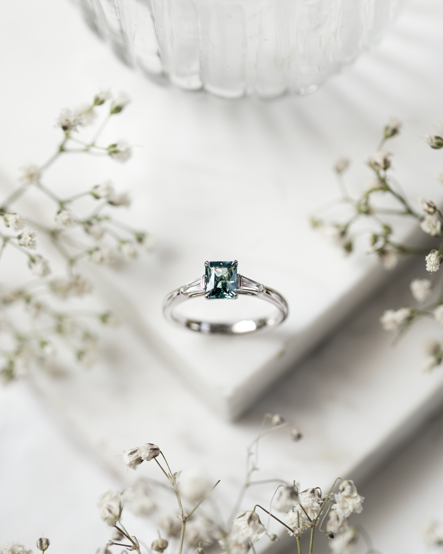 Teal Sapphire and Diamond Ring photographed on a grey marble base with white flowers