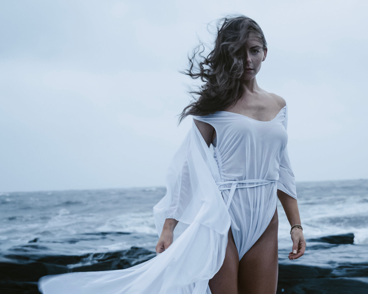 Woman in a white dress standing on a rocky beach with ocean waves.