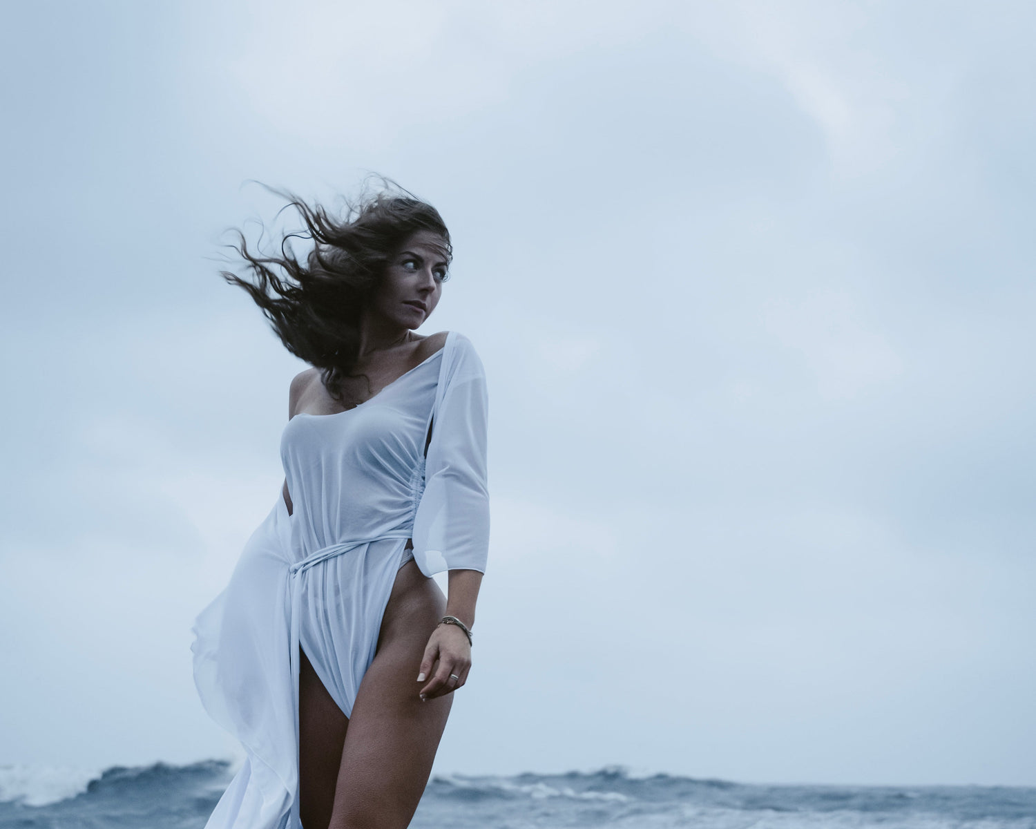 Woman in a white dress standing on a rocky beach with ocean waves in the background