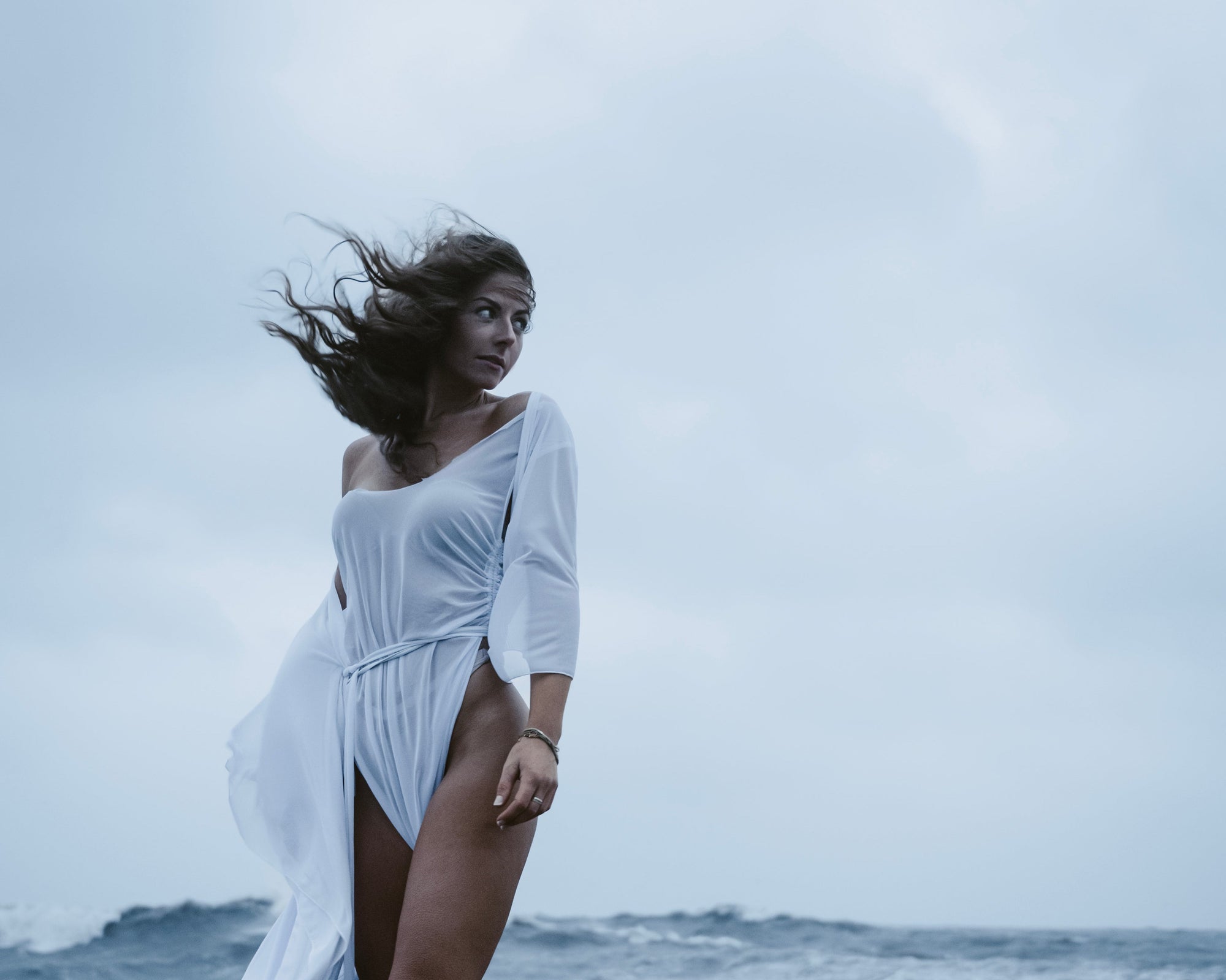 Woman in a white dress standing on a rocky beach with ocean waves in the background
