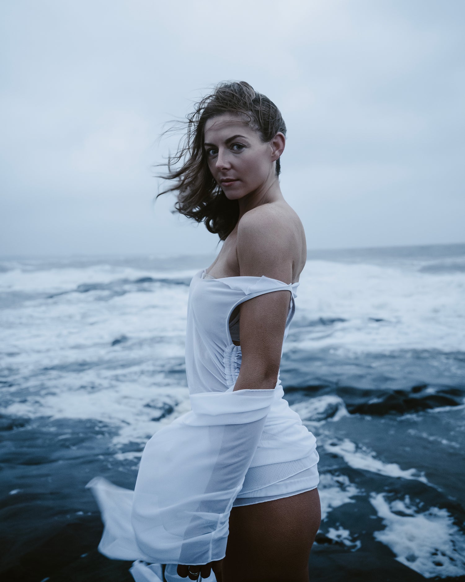 Woman in a white dress standing on a rocky shore with ocean waves in the background
